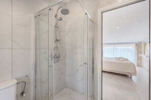 Ein modernes Badezimmer mit einer gläsernen Duschkabine, Marmorwänden und einem sichtbaren Schlafzimmer mit einem Bett.