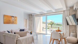 A modern living room with a sofa, dining table, and chairs, a large window with a view, and a painting on the wall.