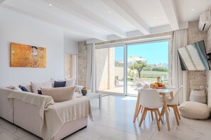 A modern living room with a sofa, dining table, and chairs, a large window with a view, and a painting on the wall.