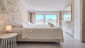 A bedroom with a stone wall, a bed with white linens, a bedside table with a lamp, and a view through a glass door.