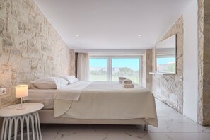 A bedroom with a stone wall, a bed with white linens, a bedside table with a lamp, and a large window with a view of greenery.