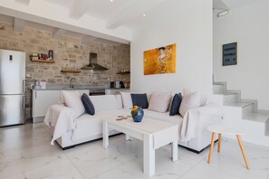 A modern living room with a white sofa, a coffee table, and a kitchen area with a stone wall and white cabinetry. - Mythos villa II,pool,Walking distance to beach (Damnoni)