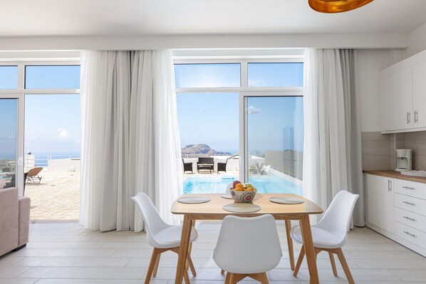 Uma cozinha moderna com armários brancos, um espaço de jantar com mesa e cadeiras, e vista para uma piscina e praia.