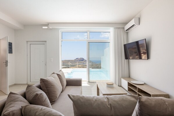 A modern living room with a sofa, a flat-screen TV, a small table, and a view of a pool and mountains.