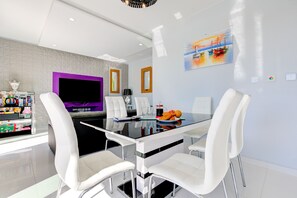 A modern dining area with a glass table, white chairs, a purple accent wall, and a painting of boats. - Villa 347 - Meo (Kouklia)