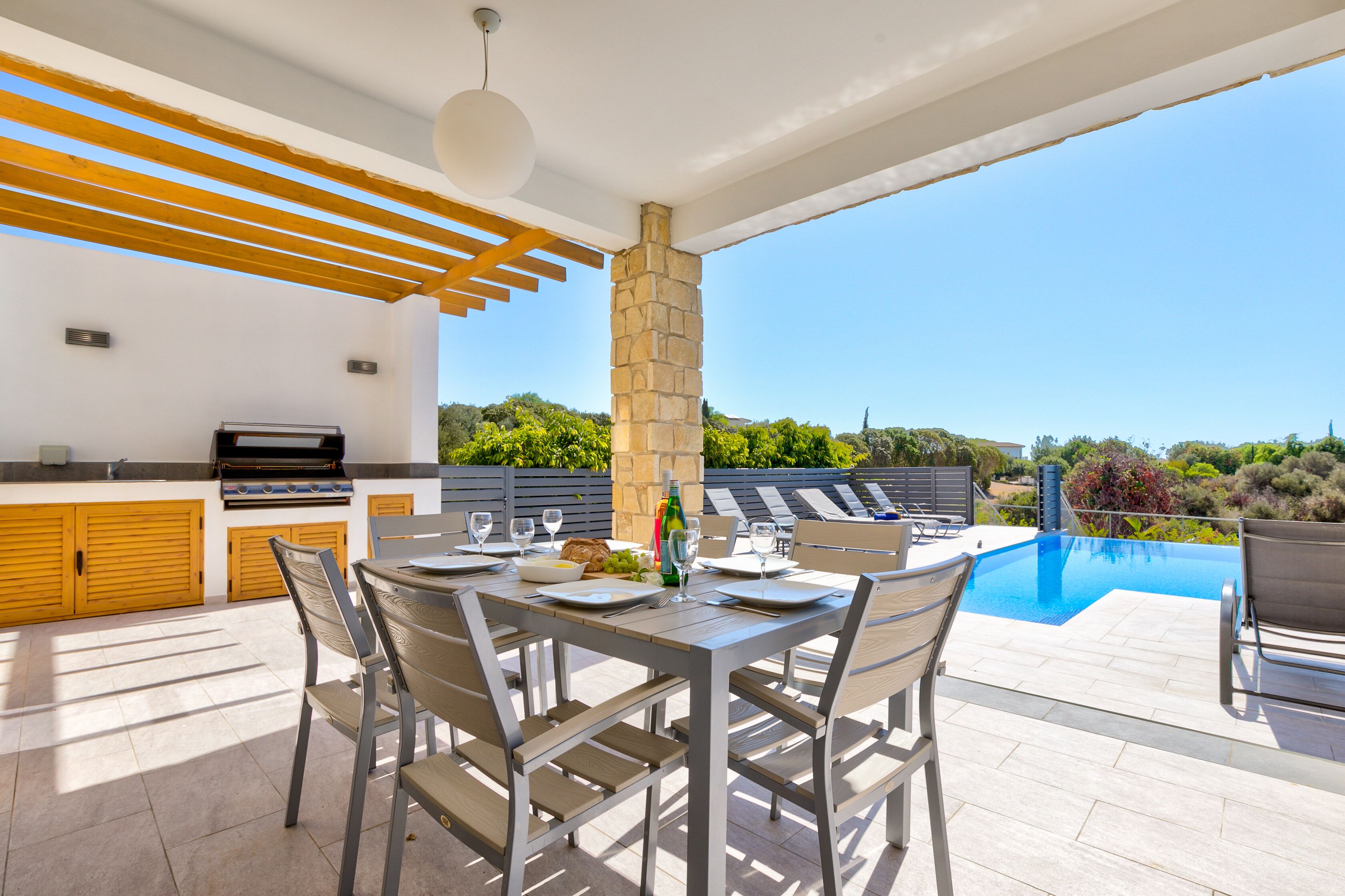 A modern outdoor dining area with a pool, stone pillar, and wooden pergola.