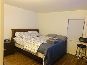 A neatly made bed with a checkered bedspread and pillows, a bedside table with a clock, and a window with a closed shutter. - Private Queen Bedroom Garvin (Sharon Hill)