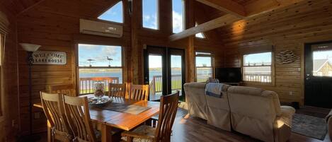 A wooden cabin interior with a dining table, chairs, a sofa, and a television.