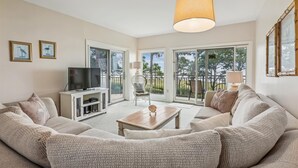 Sala de estar con un sofá, una mesa de centro, un televisor sobre un mueble bajo y una lámpara. Hay dos ventanas con vista a árboles y un balcón.