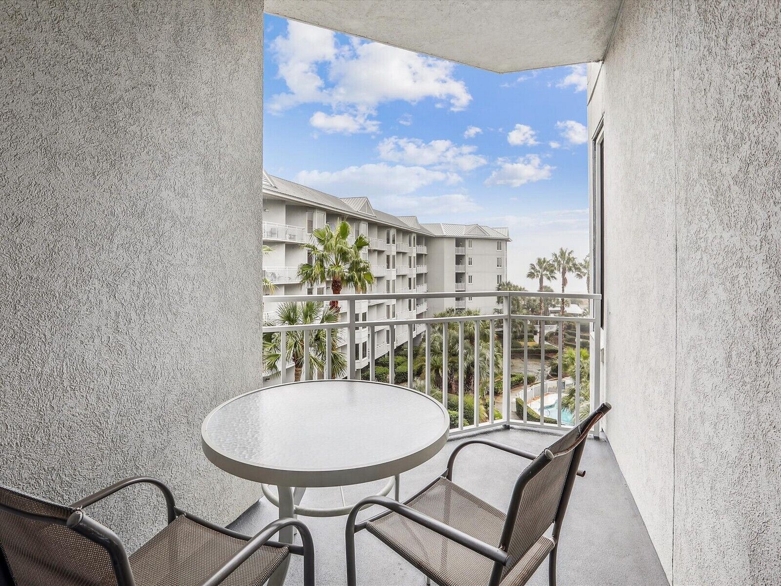 A balcony with a round table and two chairs, overlooking a building with multiple balconies and palm trees.