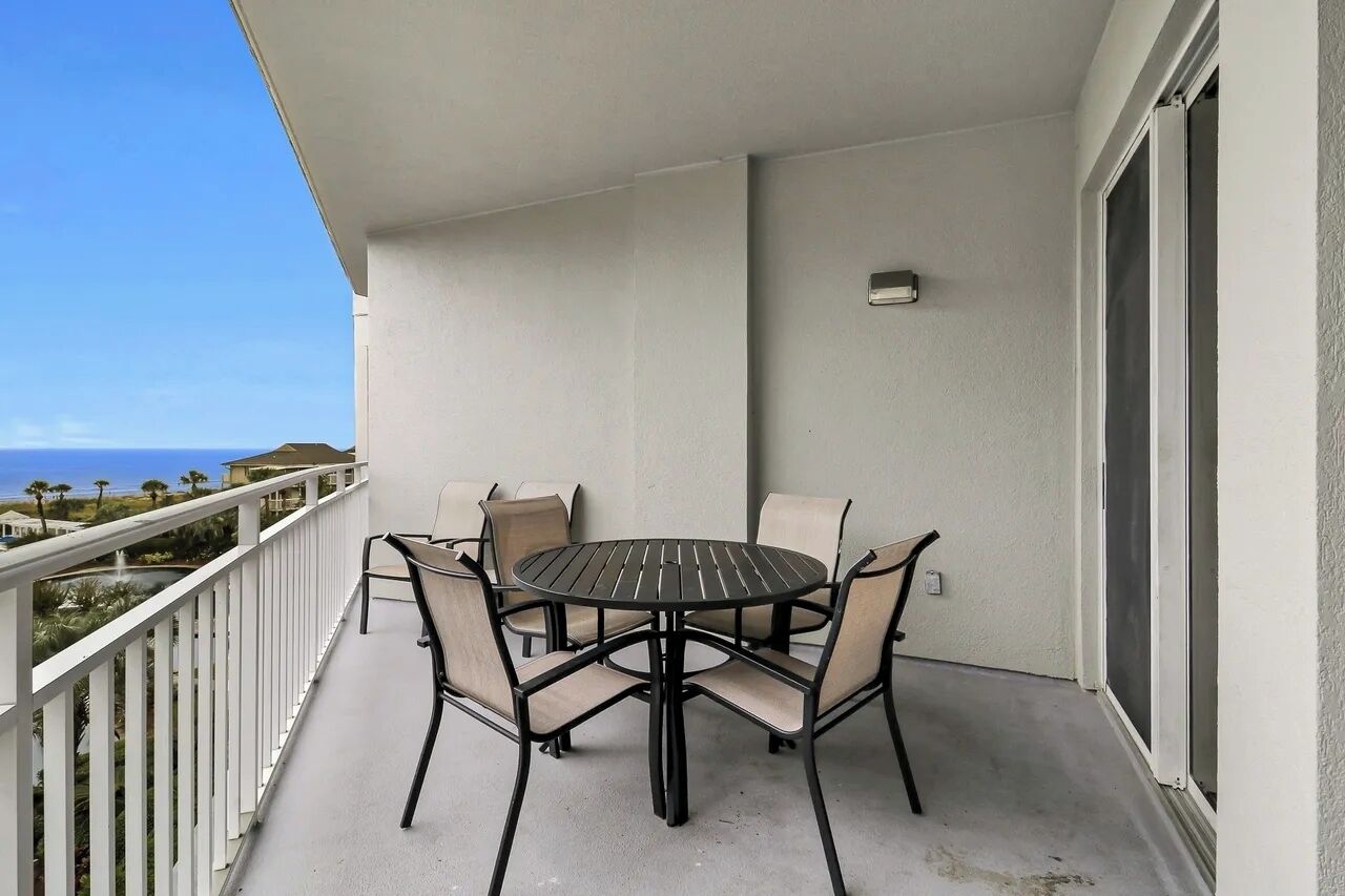 A balcony with a round table and four chairs, a white railing, a sliding glass door, and a view of the ocean and palm trees.