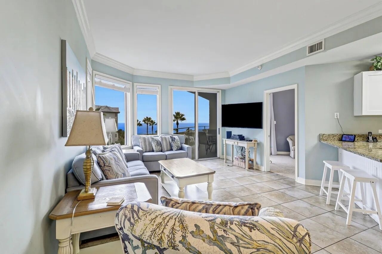 A spacious living area with a sofa, coffee table, and a TV. There are large windows offering a view of the sea.
