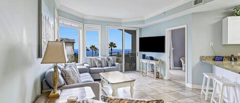A spacious living area with a sofa, coffee table, and a TV. There are large windows offering a view of the sea.
