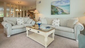 A living room with striped sofas, a glass coffee table, and a painting of a beach scene. - Enjoy the Large Wrap Around Ocean Front Balcony! (Hilton Head Island)