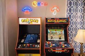 Two arcade machines, one for Pac-Man and one for Mortal Kombat, in a room with patterned wallpaper and a lamp.