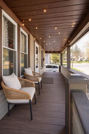 A covered porch with wicker chairs and a bench, wooden flooring, and string lights.
