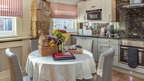 A kitchen with a stone wall, wooden beams, a dining table with a white tablecloth, and a kitchen island with a built-in oven. - 2 bedroom accommodation in Felton (Felton)