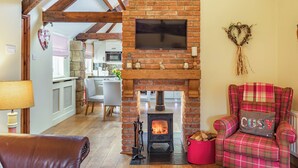 A cozy living room with a fireplace, a television mounted above it, a plaid armchair, and a dining area in the background. - 2 bedroom accommodation in Felton (Felton)