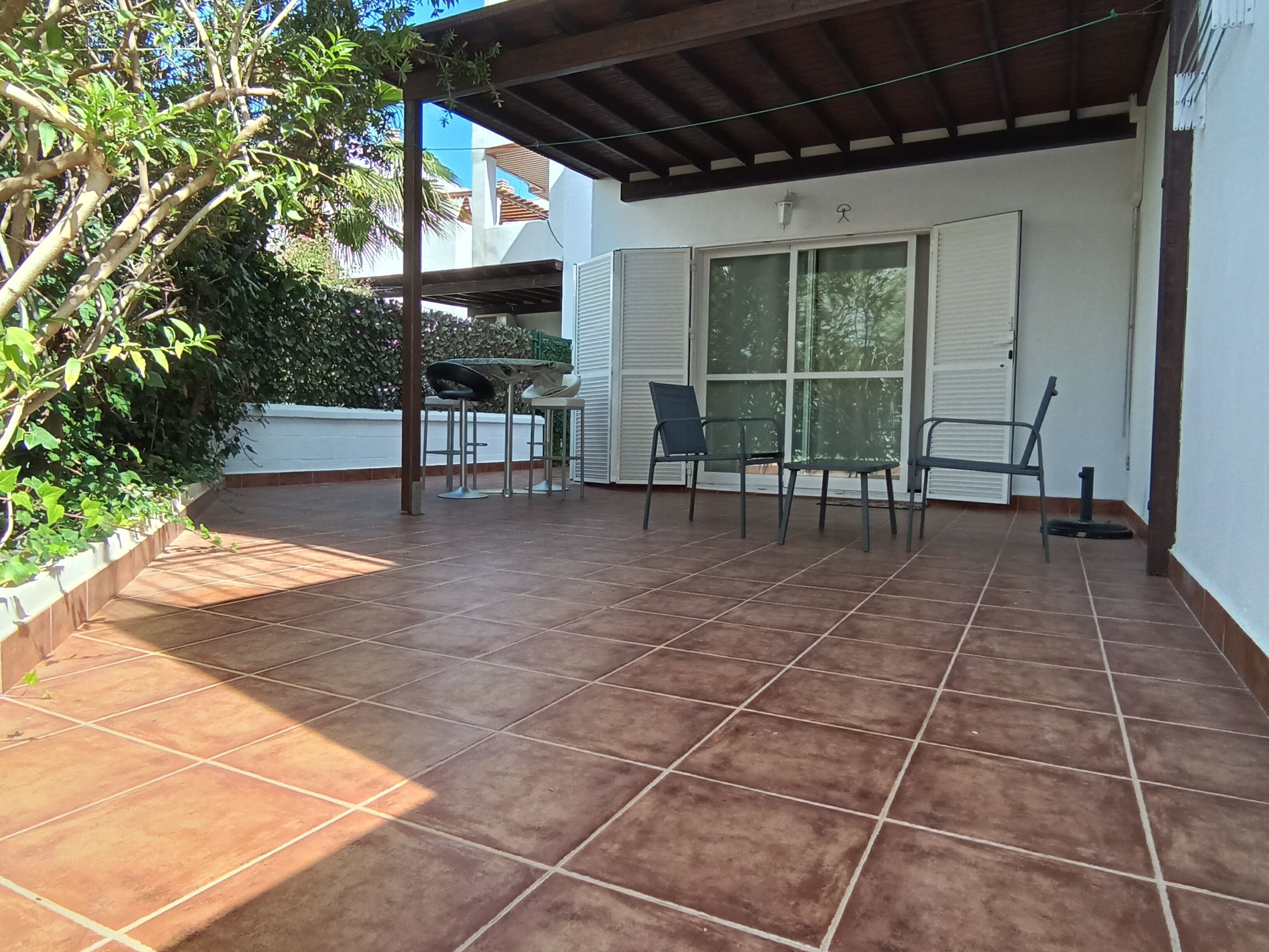 Un patio cubierto con suelo de baldosas, una pérgola y una zona de estar con sillas y una mesa.
