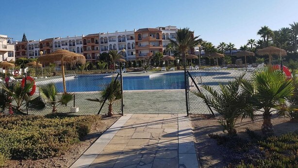 A resort pool area with a clear blue pool, palm trees, and a row of multi-story buildings in the background. - NATURIST DUPLEX IN FIRST LINE OF VERA PLAYA! (Vera)