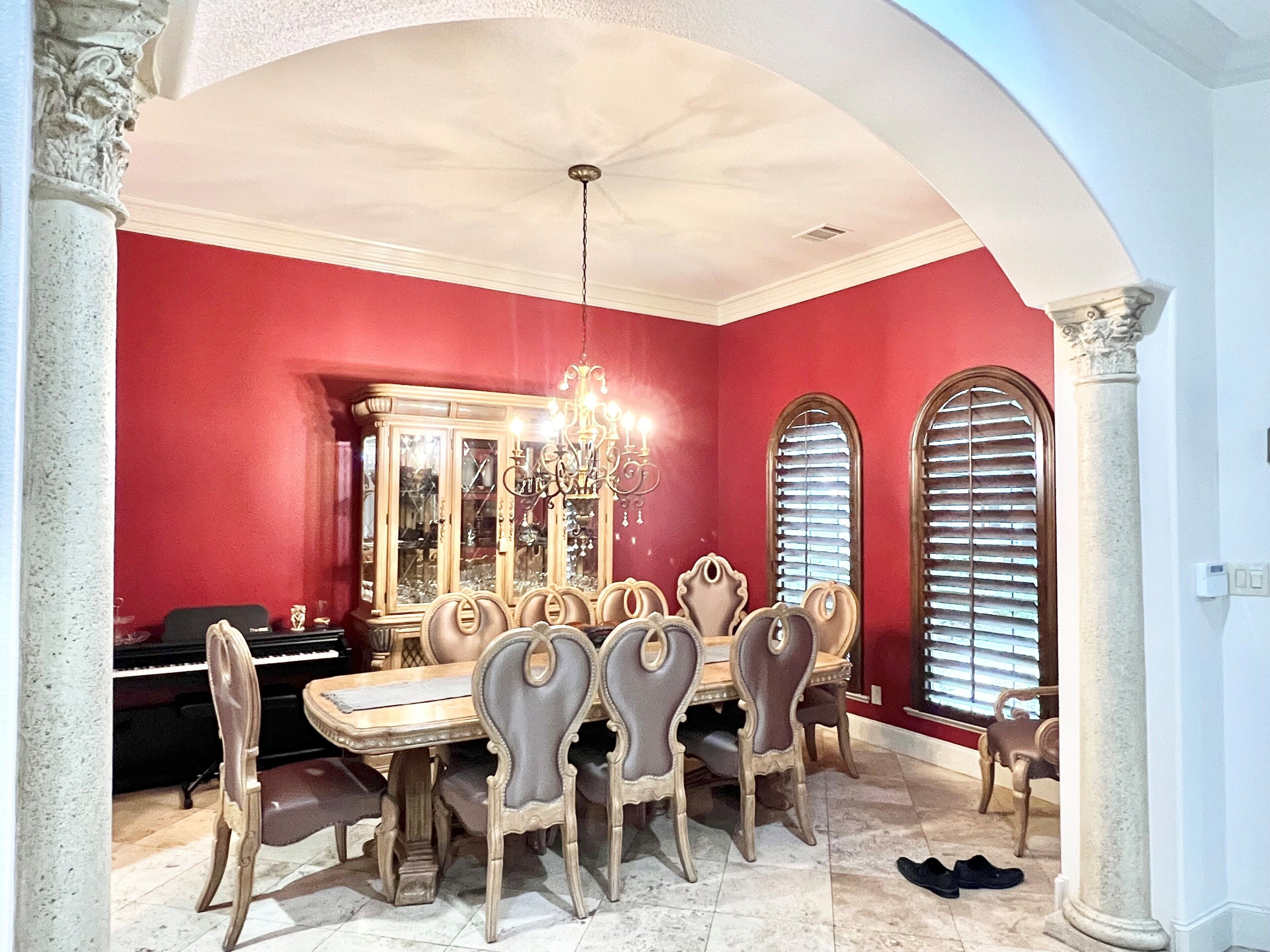 A dining room with a large wooden table and eight chairs, a chandelier, and a piano in the background.