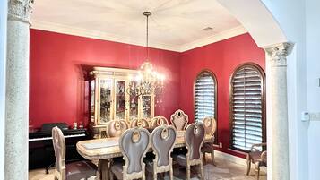 A dining room with a large wooden table and eight chairs, a chandelier, and a piano in the background.
