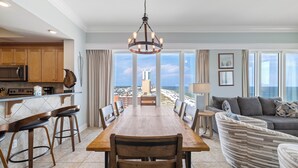 Een ruime eethoek met een grote houten tafel en stoelen, een keuken met houten kastjes, en uitzicht op het strand en de skyline door grote ramen.