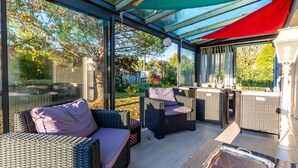 A covered outdoor seating area with wicker furniture and glass walls. - Gawa's House - Maison avec jardin proche mer (Meschers-sur-Gironde)