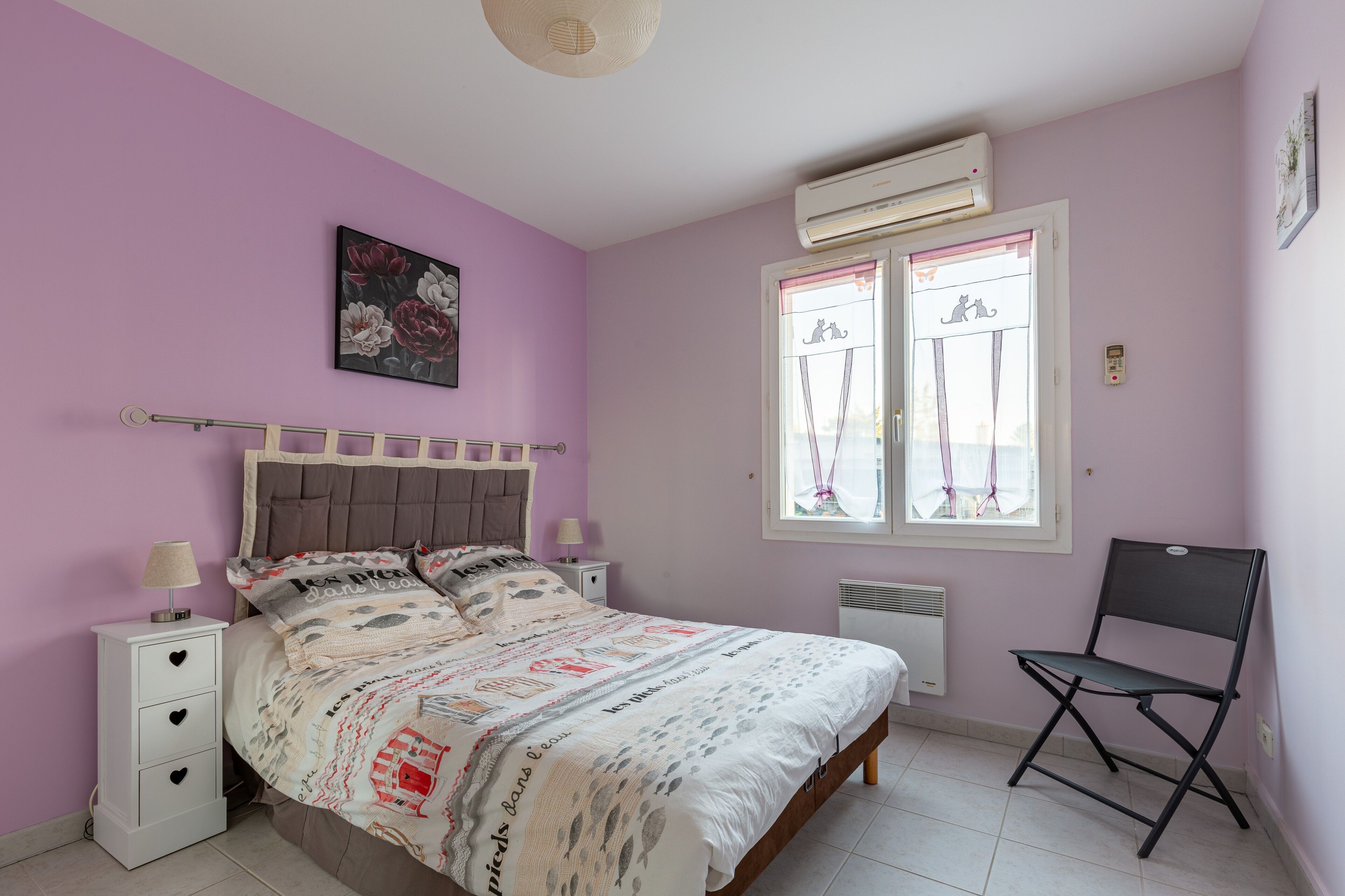 Ein Schlafzimmer mit einem Bett, einem Nachttisch, einem Sessel, einem Fenster mit Vorhängen und einer Klimaanlage.