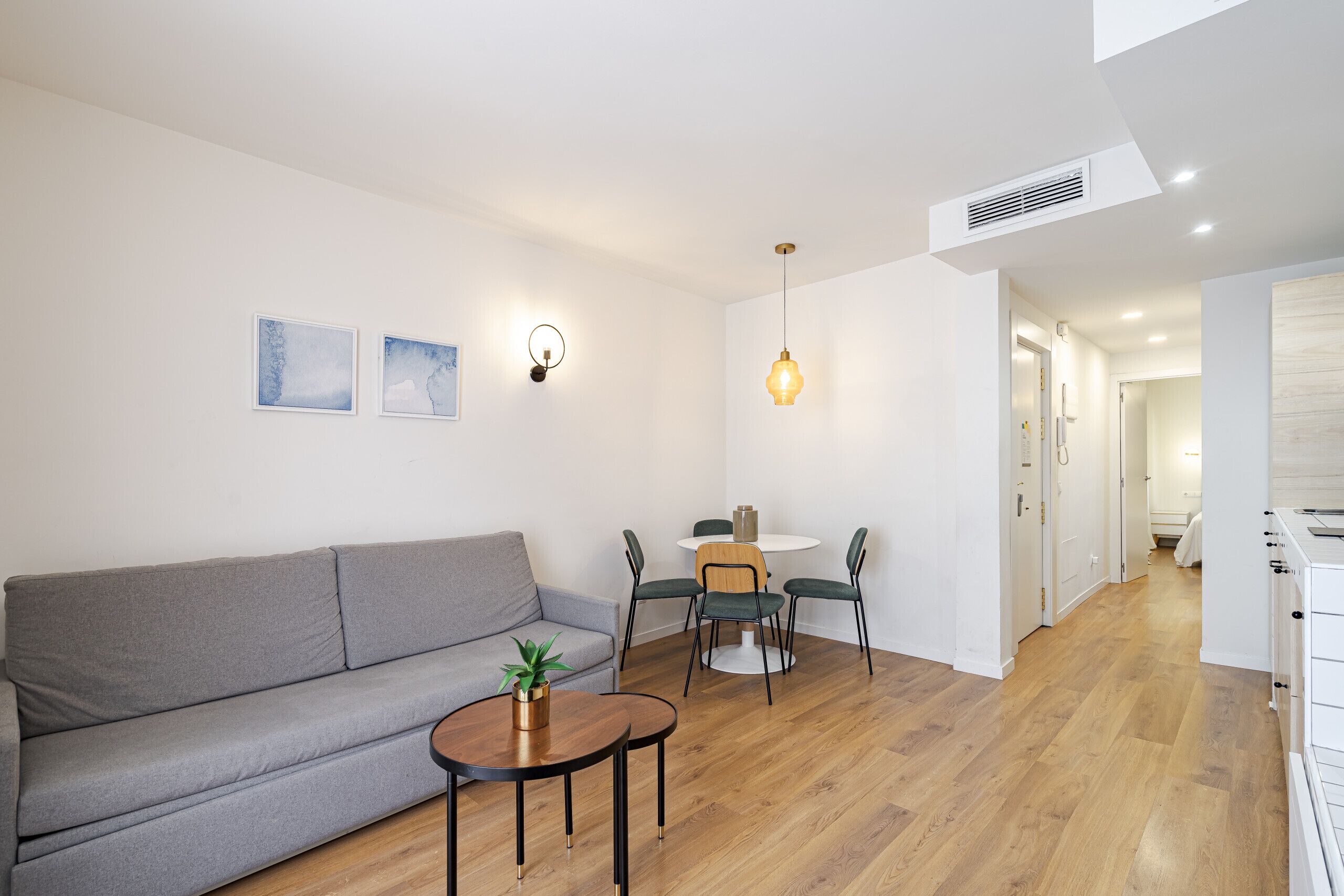 Ein modernes Wohnzimmer mit grauem Sofa, einem kleinen runden Tisch mit Pflanze, einem Essbereich mit weißem Tisch und vier Stühlen sowie einem Flur, der in einen Küchenbereich führt.