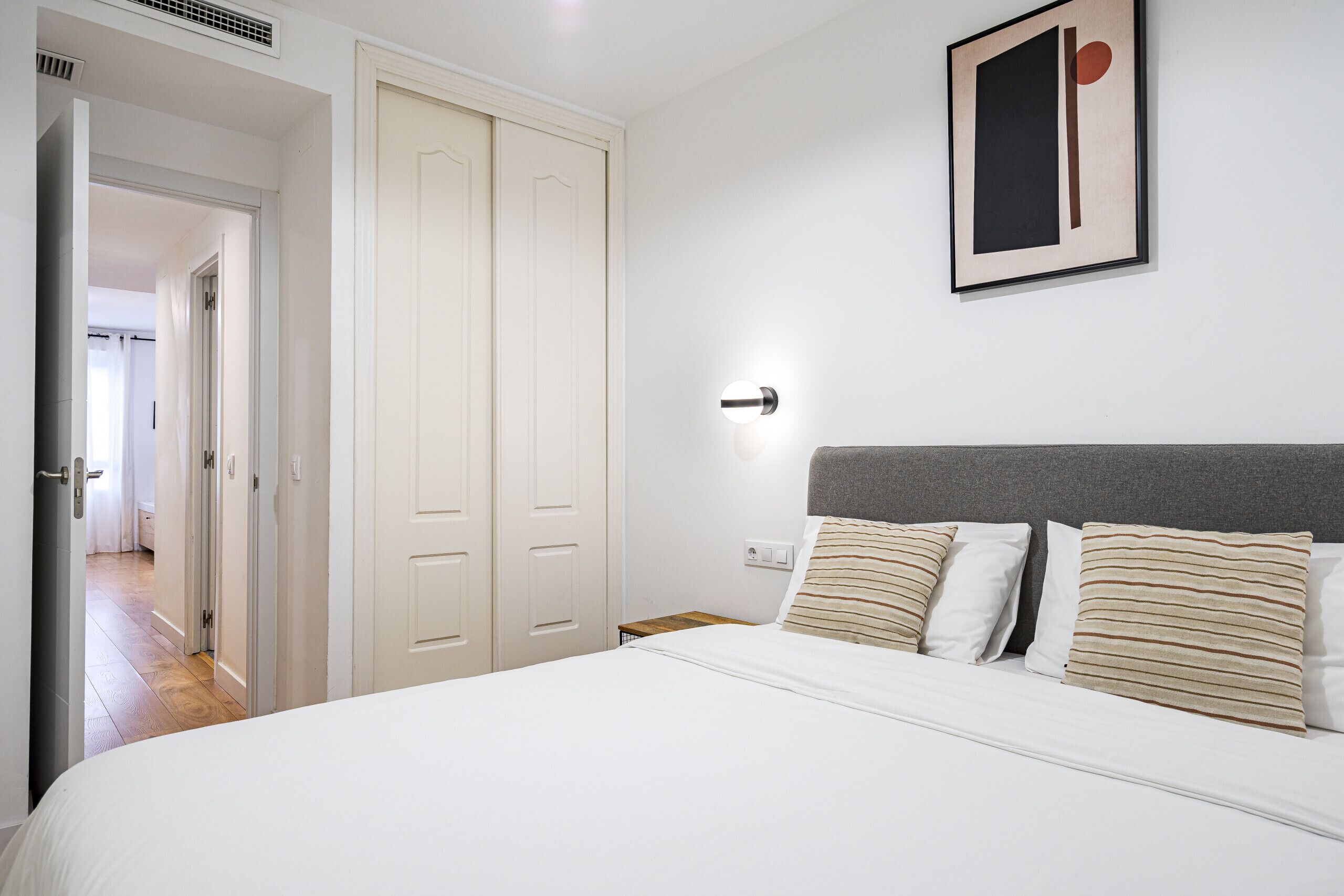 A bedroom with a large bed, a bedside table, a nightstand, a framed picture on the wall, and a door leading to another room.