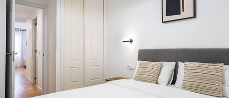 A bedroom with a large bed, a bedside table, a nightstand, a framed picture on the wall, and a door leading to another room.