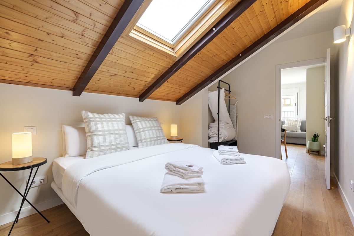 A bedroom with a skylight, a bed with white linens, a bedside table with a lamp, and a hanging rack with towels.