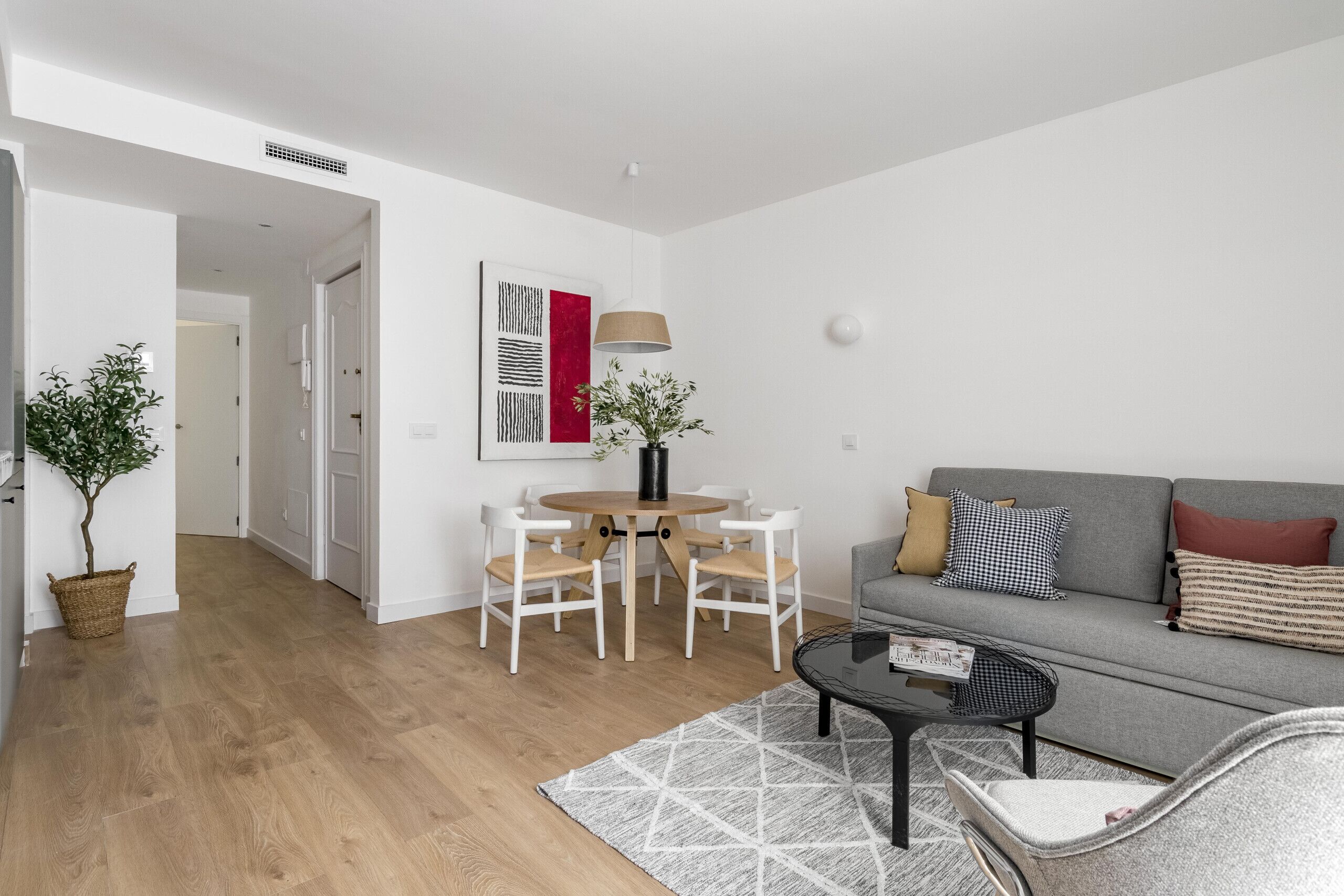 A modern living room with a grey sofa, a round wooden table with chairs, a coffee table, and a potted plant.