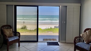 Een kamer met een grote schuifglazen deur die uitkomt op uitzicht op het strand. Er staan twee rieten stoelen met kussens.