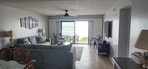 A living room with a sofa, chairs, a coffee table, and a TV. There is a sliding glass door leading to a balcony with a view of the ocean.