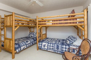 A room with two bunk beds, each with a checkered quilt, a wooden chair, and a ceiling fan.