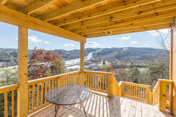 A wooden deck with a metal table and chairs, overlooking a valley with autumn foliage and distant buildings.