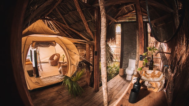 A rustic, thatched-roof tent with a wooden floor, a wicker chair, and a small table with a plant. - Uman Glamping & Cenote Tulum (Tulum)