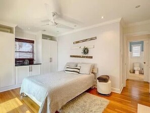 Une chambre à coucher avec un lit, un ventilateur de plafond blanc, une petite table de chevet et une fenêtre avec des stores.
