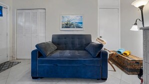 Ein blaues Sofa mit Kissen, ein gerahmtes Bild von einem Boot, eine Lampe und ein Korbwäscher mit Gegenständen.