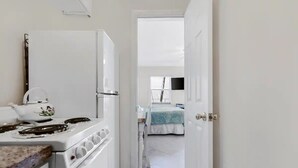 Ein schmaler Flur führt zu einem Schlafzimmer mit Bett und Fenster. In der Küchenzeile ist ein weißer Herd mit Wasserkocher zu sehen.