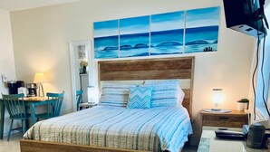 Ein Schlafzimmer mit einem hölzernen Bett, einem Schreibtisch mit Stühlen, einer Lampe und einem Gemälde vom Meer an der Wand.