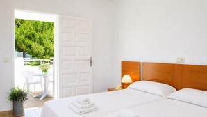 A hotel room with two beds, a wooden headboard, a bedside table with a lamp, a small potted plant, and a view of a white door leading to an outdoor area with a table and chairs. - Villa Roca Nova - Four Bedroom Villa, Sleeps 8 (Cala d'Or)