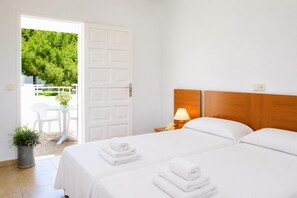 A hotel room with two beds, a wooden headboard, a bedside table with a lamp, a small potted plant, and a view of a white door leading to an outdoor area with a table and chairs. - Villa Roca Nova - Four Bedroom Villa, Sleeps 8 (Cala d'Or)