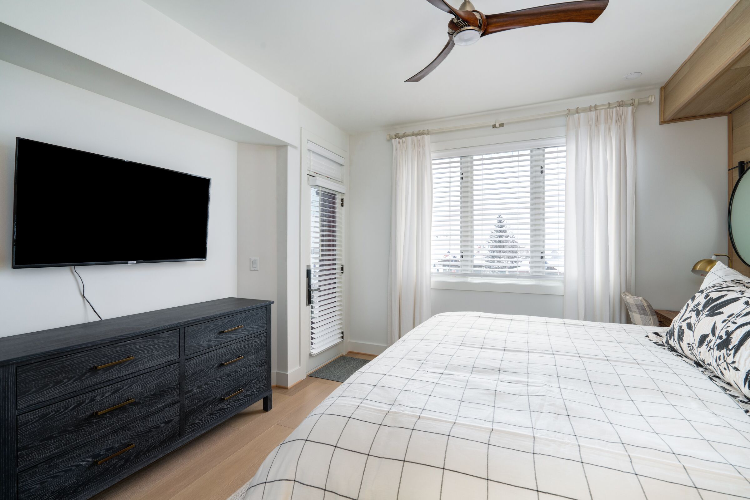 A bedroom with a bed, a TV mounted on the wall, a ceiling fan, and a window with blinds.