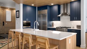 A modern kitchen with dark blue cabinets, a large island with bar stools, stainless steel appliances, and a wooden ceiling.