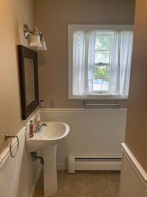 Un baño con lavamanos blanco, espejo, ventana con cortinas blancas y un perchero para toallas.