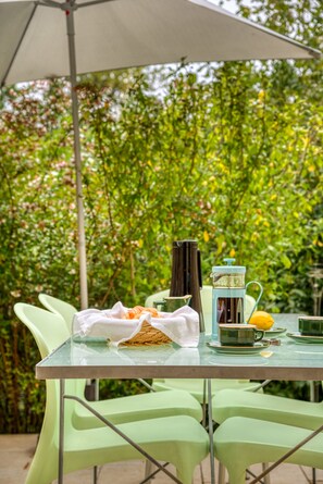 A table with a clear glass top, surrounded by green chairs, under a white umbrella, set against a backdrop of lush greenery. - Gîte-Deluxe-Countryside view-Ensuite (Servies-en-Val)