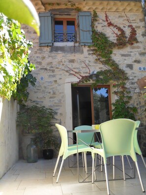 A stone building with a window and shutters, a door, and a table with chairs. - Gîte-Deluxe-Countryside view-Ensuite (Servies-en-Val)